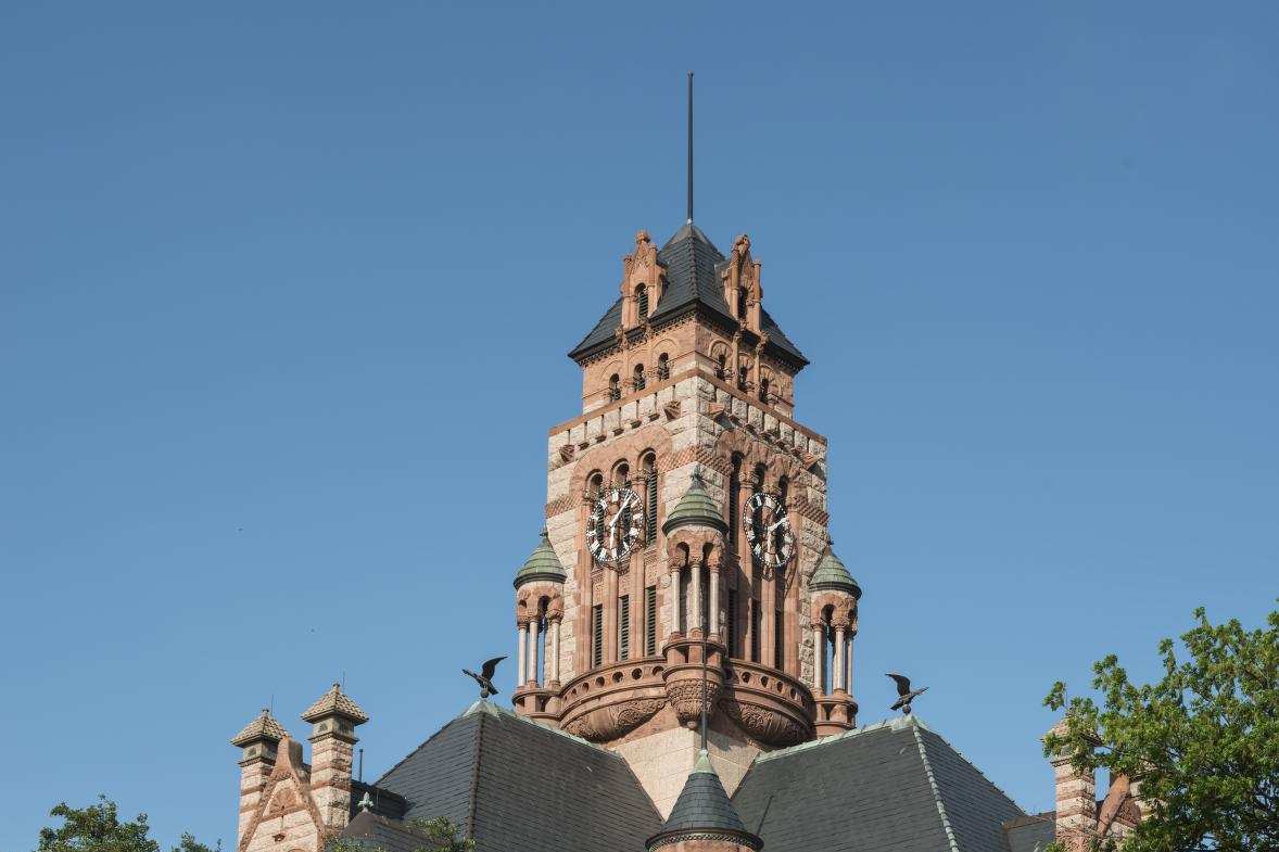 Color photograph of a red and cream stone courthouse tower with two clock faces, small open turrets and a steep slate roof rising above leafy trees against a clear blue sky, with a red traffic sign in the foreground.