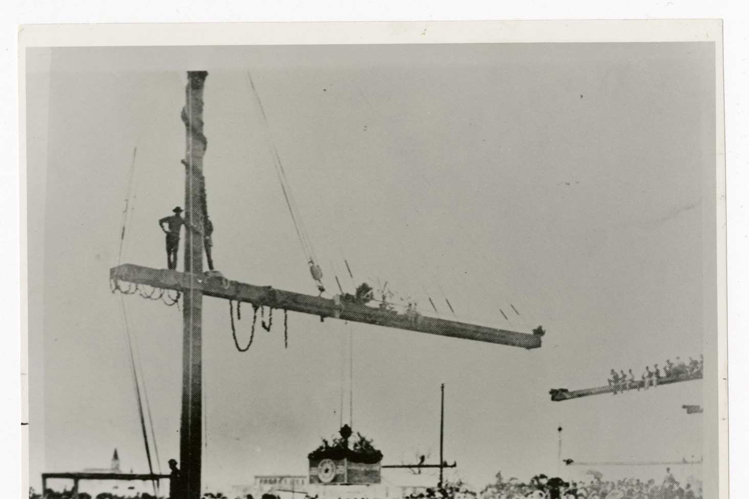 Black and white photograph of a dense crowd of men in hats around a tall timber derrick with a long boom, several men standing on the boom, a draped stand with a star emblem in the middle distance and a flag bearing a single star.