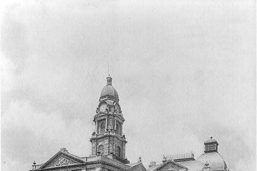Black and white view of a large stone courthouse with columned porches, a central clock tower under a small dome, a lawn with young trees and horse drawn wagons on the street in front.