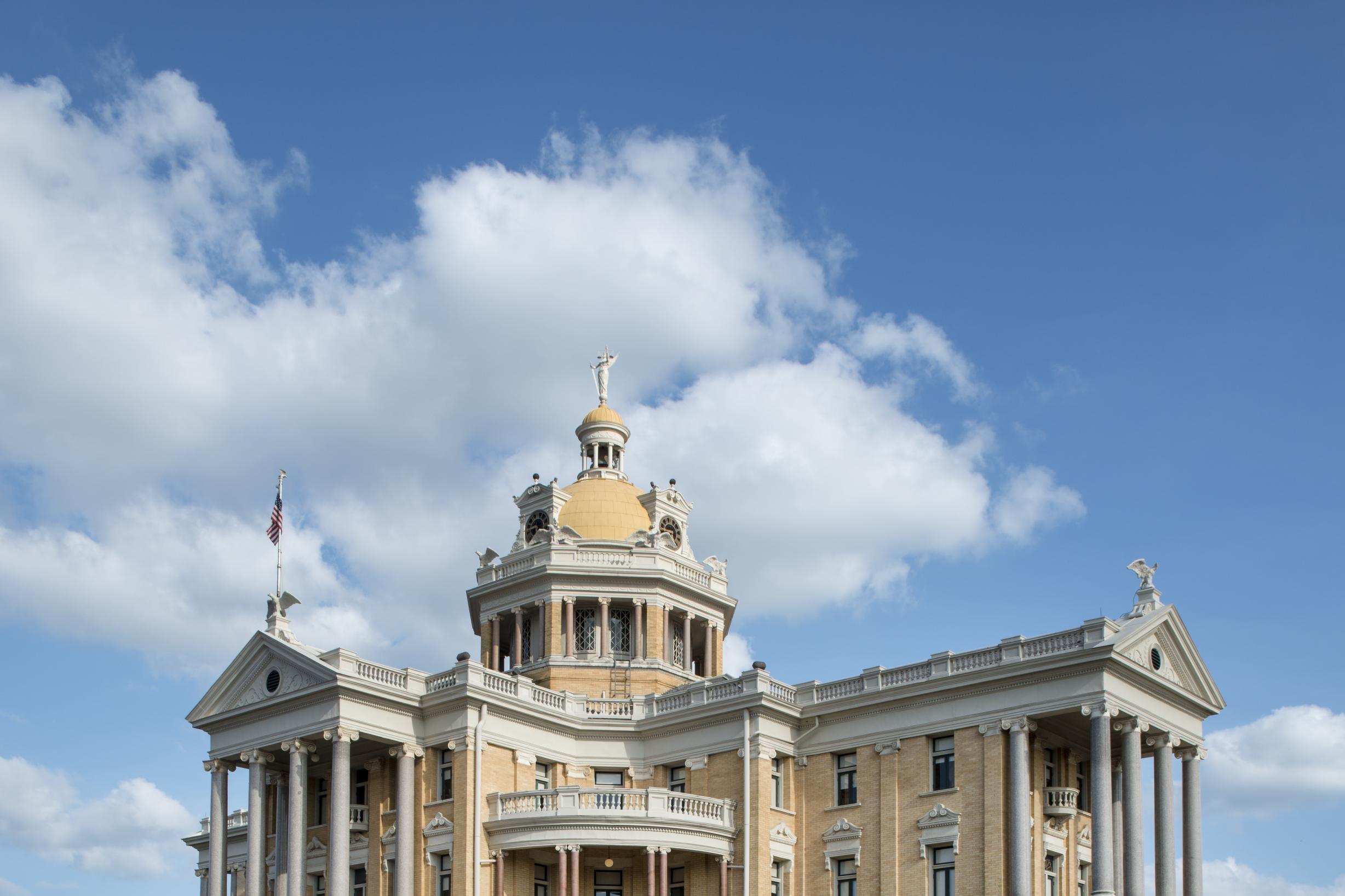 Color photograph of a pale yellow brick courthouse with tall gray columns on its porches, a domed cupola topped by a statue, and a low brick wall along the street under a blue sky with clouds.