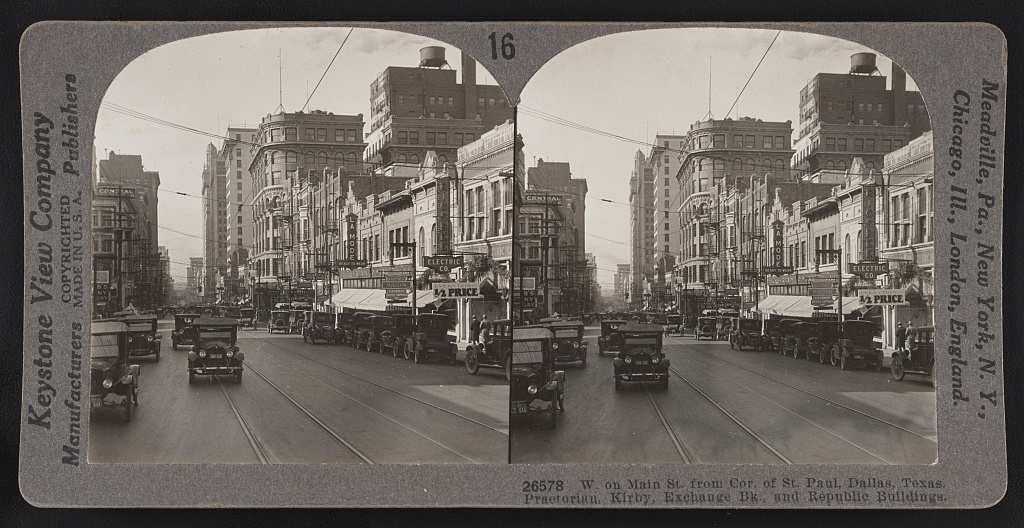A stereograph card with twin photographs looking down Main Street in Dallas, with tall commercial buildings, parked automobiles, streetcar tracks and shop signs, and a printed caption along the bottom.