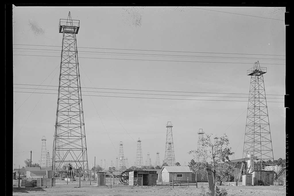 Two tall steel oil derricks stand over a flat yard of sheds, a bull wheel and a small tree, with a line of more derricks across the horizon beneath power lines.