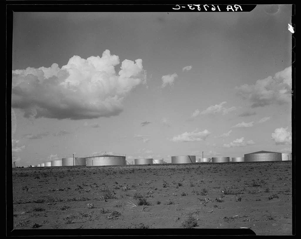 A row of low white oil storage tanks stretches along the horizon of a flat, sparsely vegetated field under a wide sky with large cumulus clouds.