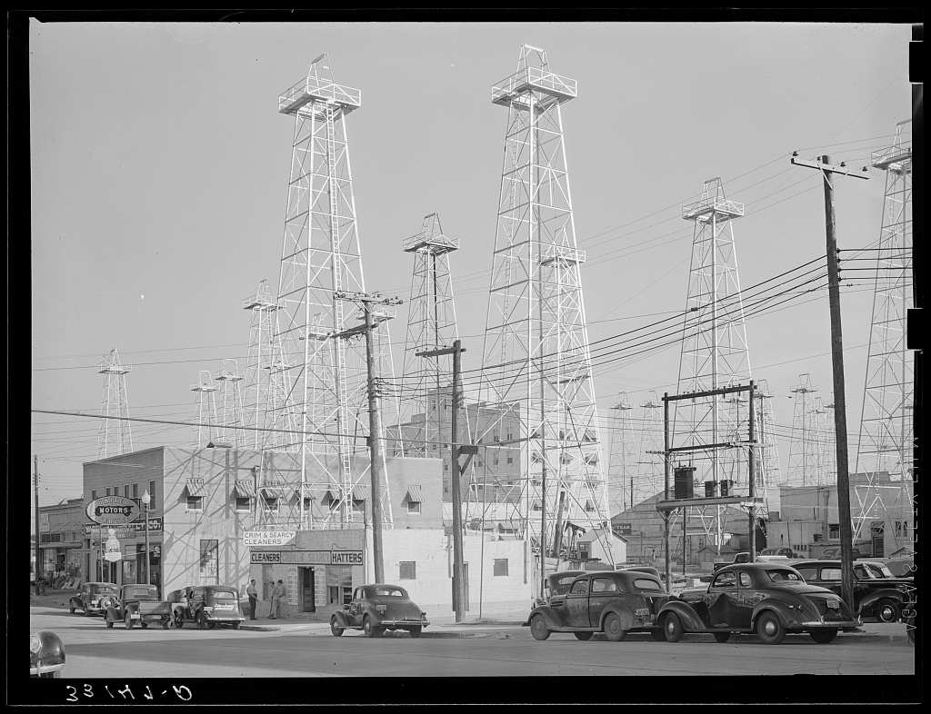 A street corner in Kilgore with a cleaners and hatters shop and parked cars of the late 1930s, beneath tall steel oil derricks standing among the downtown buildings.
