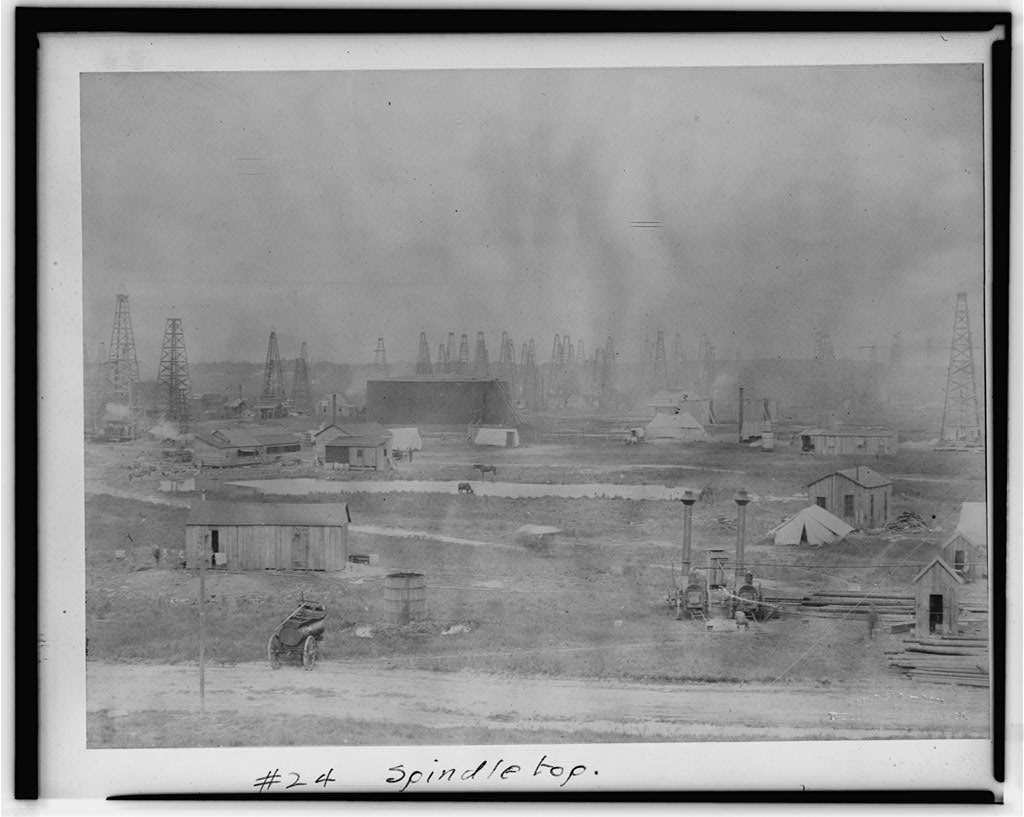 A wide field of wooden oil derricks on flat ground behind rough plank sheds, tents, a round tank and a horse drawn cart, with the handwritten caption number 24 Spindletop.