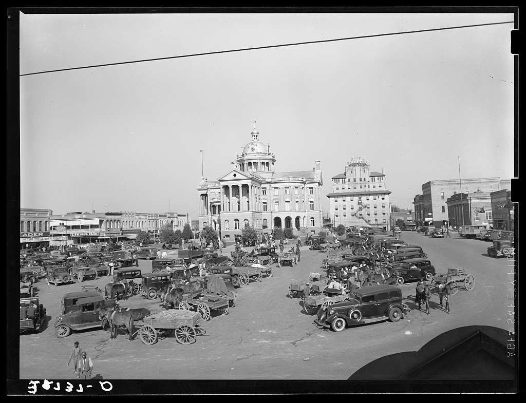 Black and white photograph of a town square crowded with parked cars, mule drawn wagons and people, with a domed and columned courthouse in the center background and brick commercial buildings on both sides.