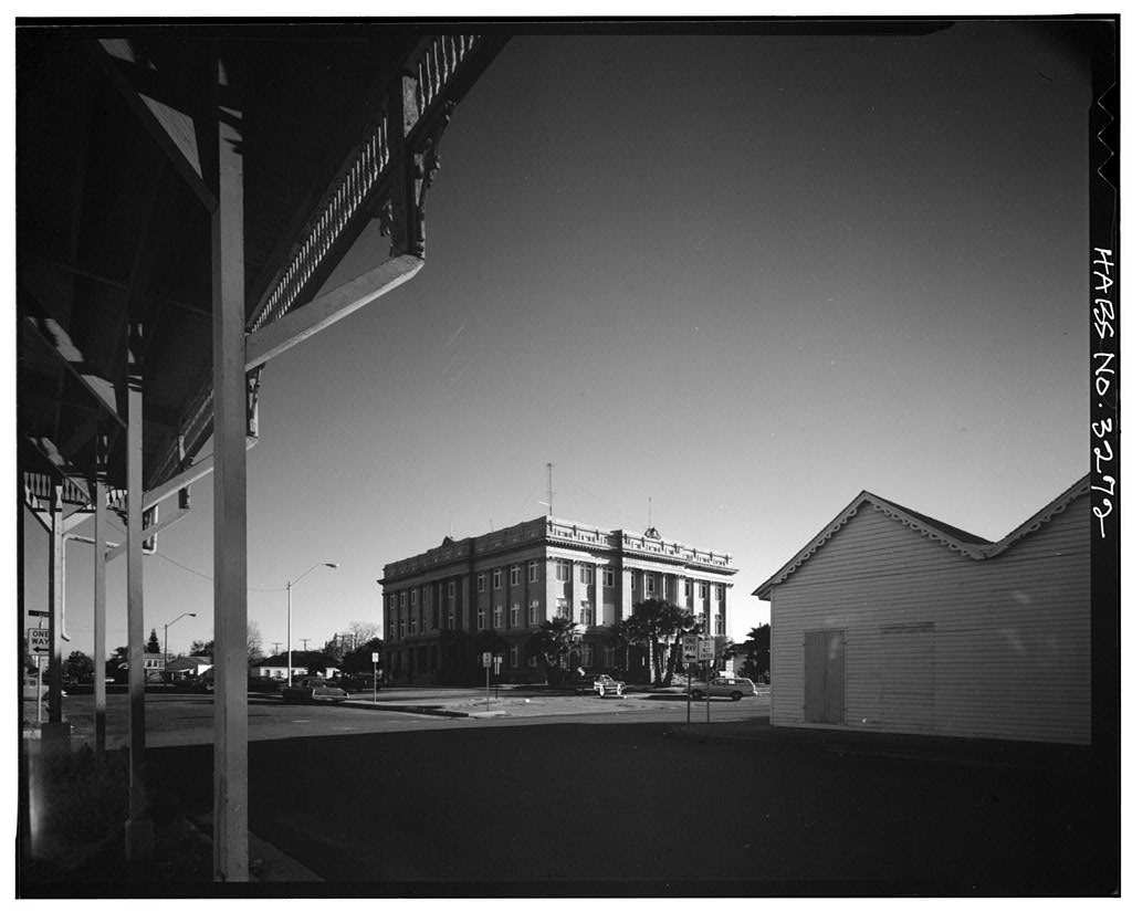 Black and white photograph looking from under the bracketed porch of a wooden building across an empty street to a large four story courthouse with a columned upper facade and palm trees at its base.