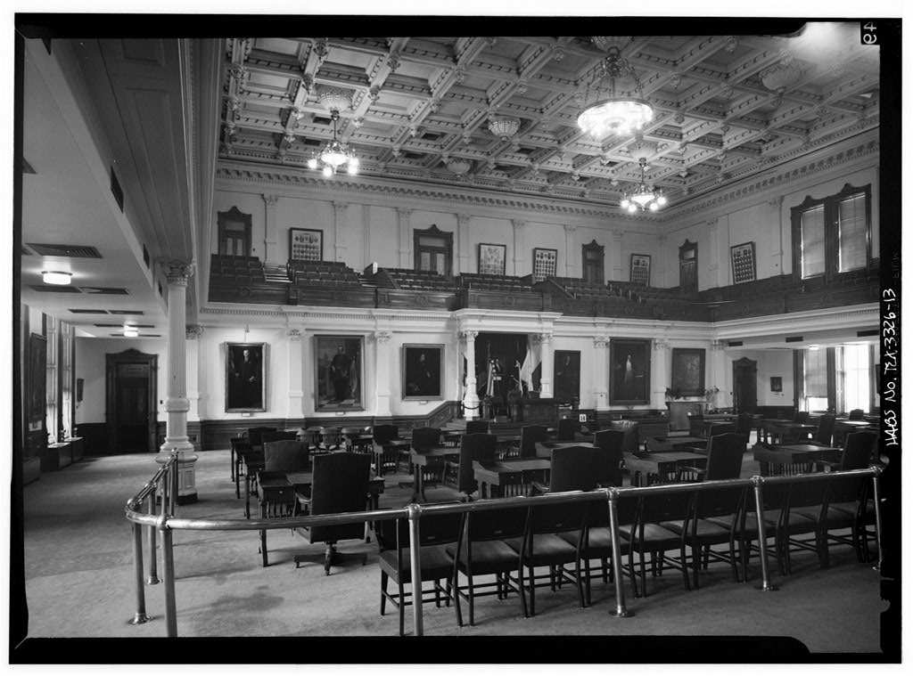 A black and white photograph of the Texas Senate chamber, with rows of wooden desks and chairs behind a brass rail, portraits on the walls, a gallery above and chandeliers hanging from a coffered ceiling.