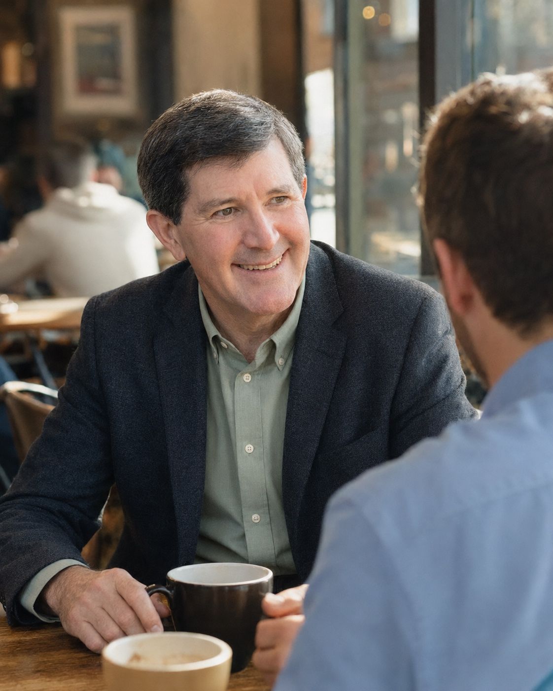 Smiling across a cafe table with a coffee cup in hand, another person seen from behind in the foreground